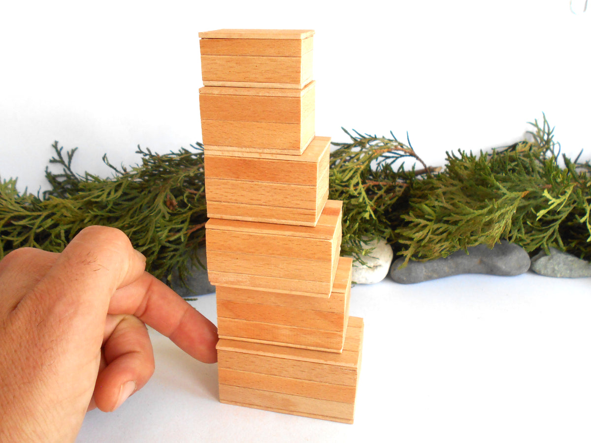 A hand is balancing a stack of small, light-colored wooden miniature boxes on a white surface, with green foliage and small stones in the background.