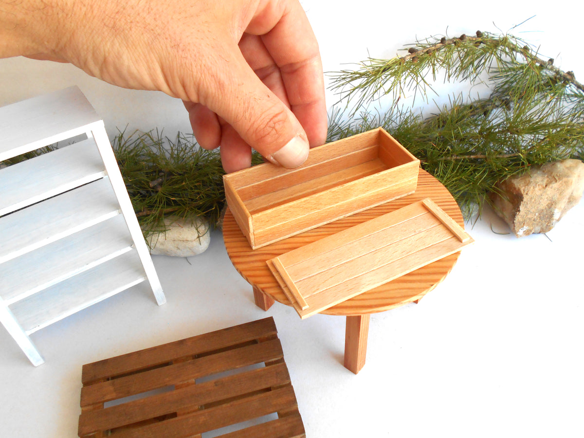 A hand holding a miniature light wood box next to a matching small table and tray, with a miniature white shelf and greenery in the background.