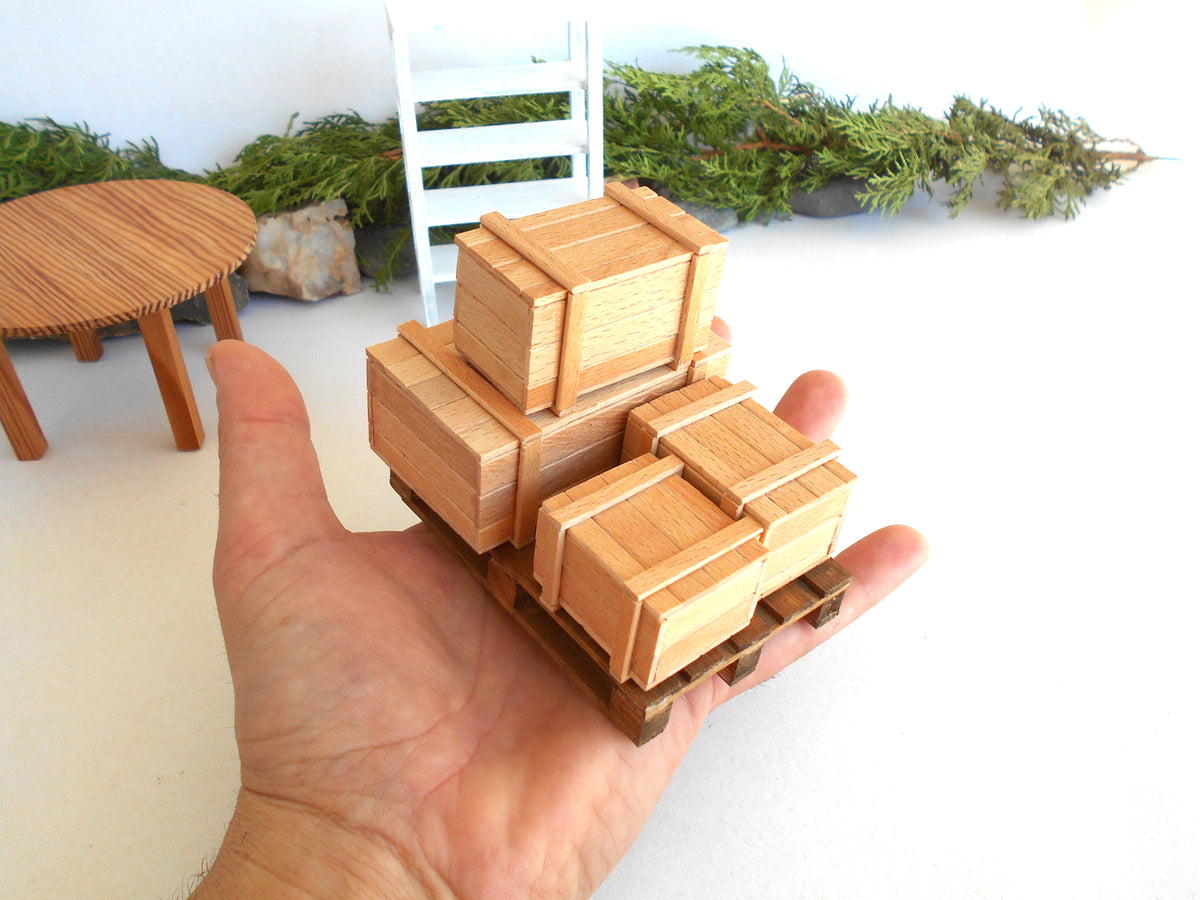 A miniature wooden crate set on a small wooden pallet, held in a person's hand to show scale, with a miniature white chair and round table in the background.