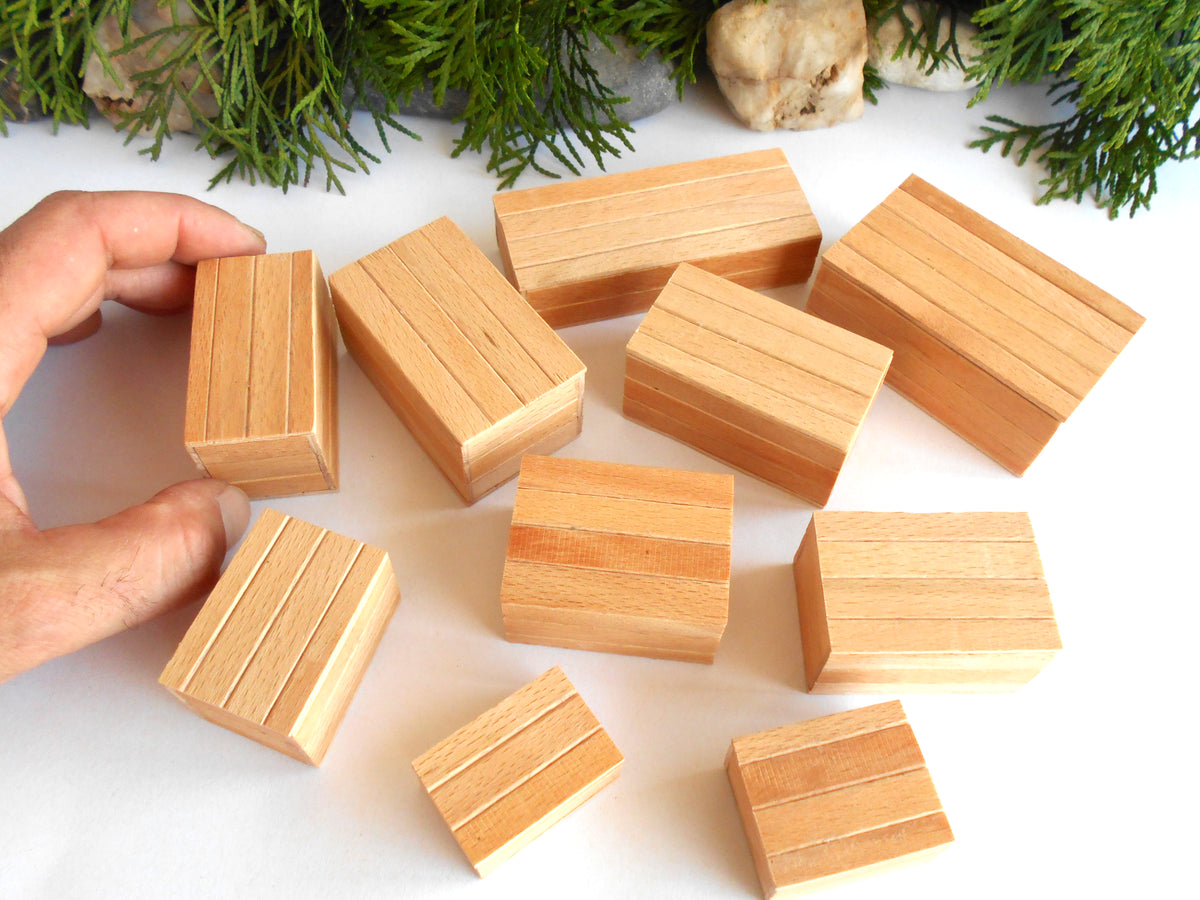 A hand is holding one of several small rectangular wooden boxes with grooved sides, arranged on a white surface with green foliage in the background.