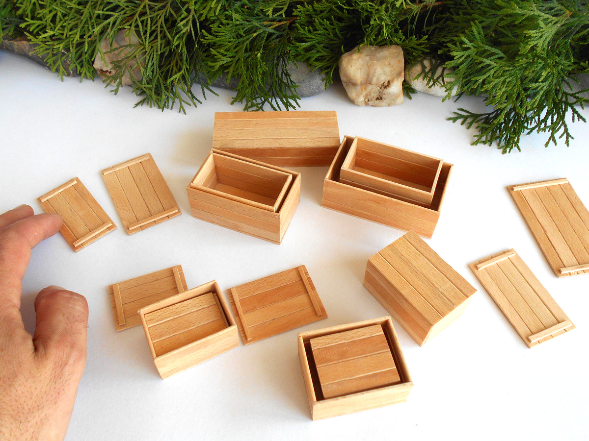 A set of miniature light wood boxes and lids, including a small wooden hand holding one piece on the left, arranged on a white surface with green pine branches in the background.