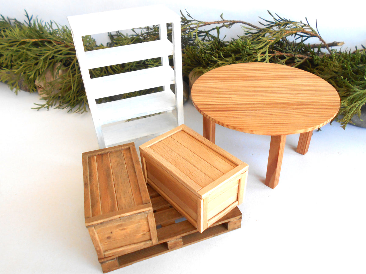 A miniature dollhouse furniture set featuring a round light wood table, two wooden storage boxes on a small wooden base, and a white slatted shelf, styled with green pine branches on a white background.