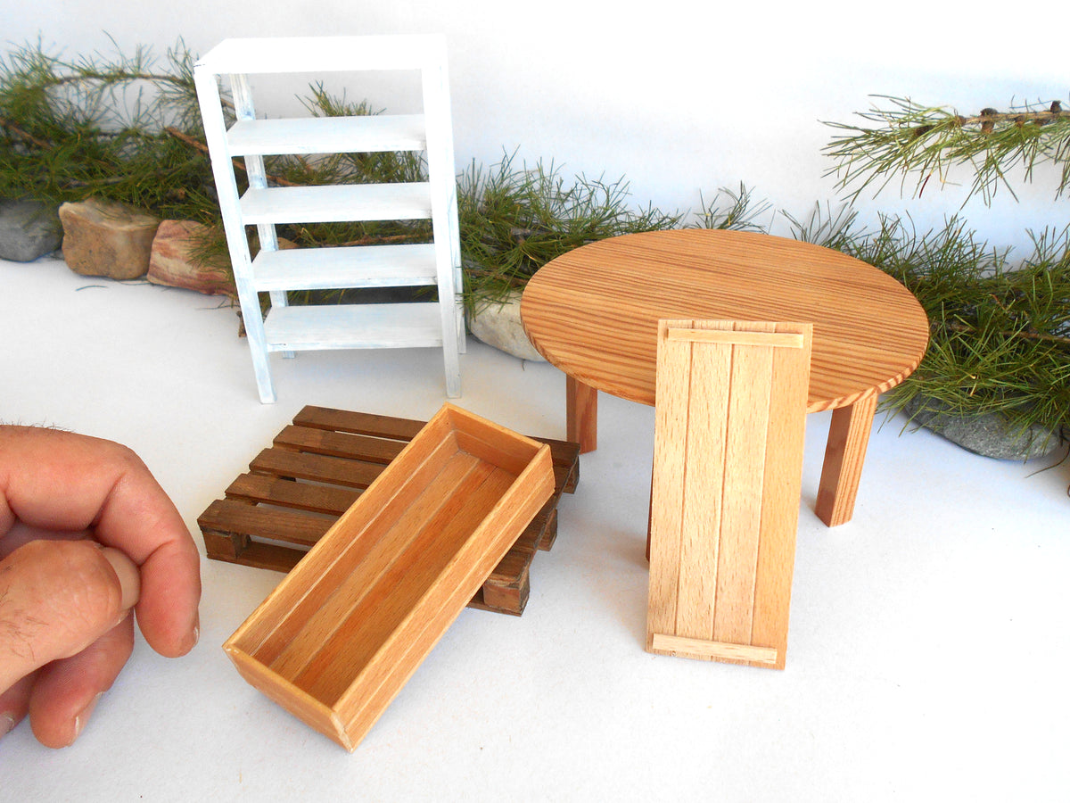 A miniature dollhouse furniture set including a round wooden table, a rectangular wooden tray, a wooden cabinet, a wooden shelf, and a wooden crate, shown with a hand for scale against a white background with faux greenery.