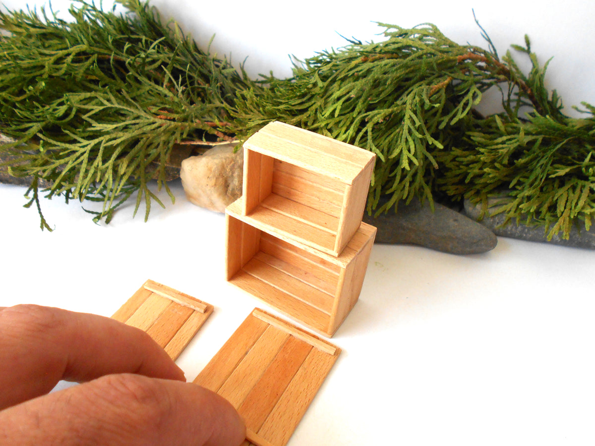 A close-up of a hand assembling miniature light wood storage boxes or shelves, with green foliage and small stones in the background on a white surface.
