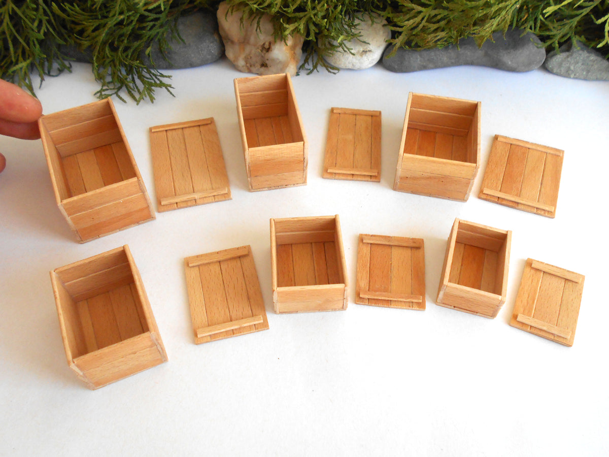 A set of miniature light wood crates and boxes arranged on a white surface, with a hand holding one crate on the left and decorative greenery and stones in the background.
