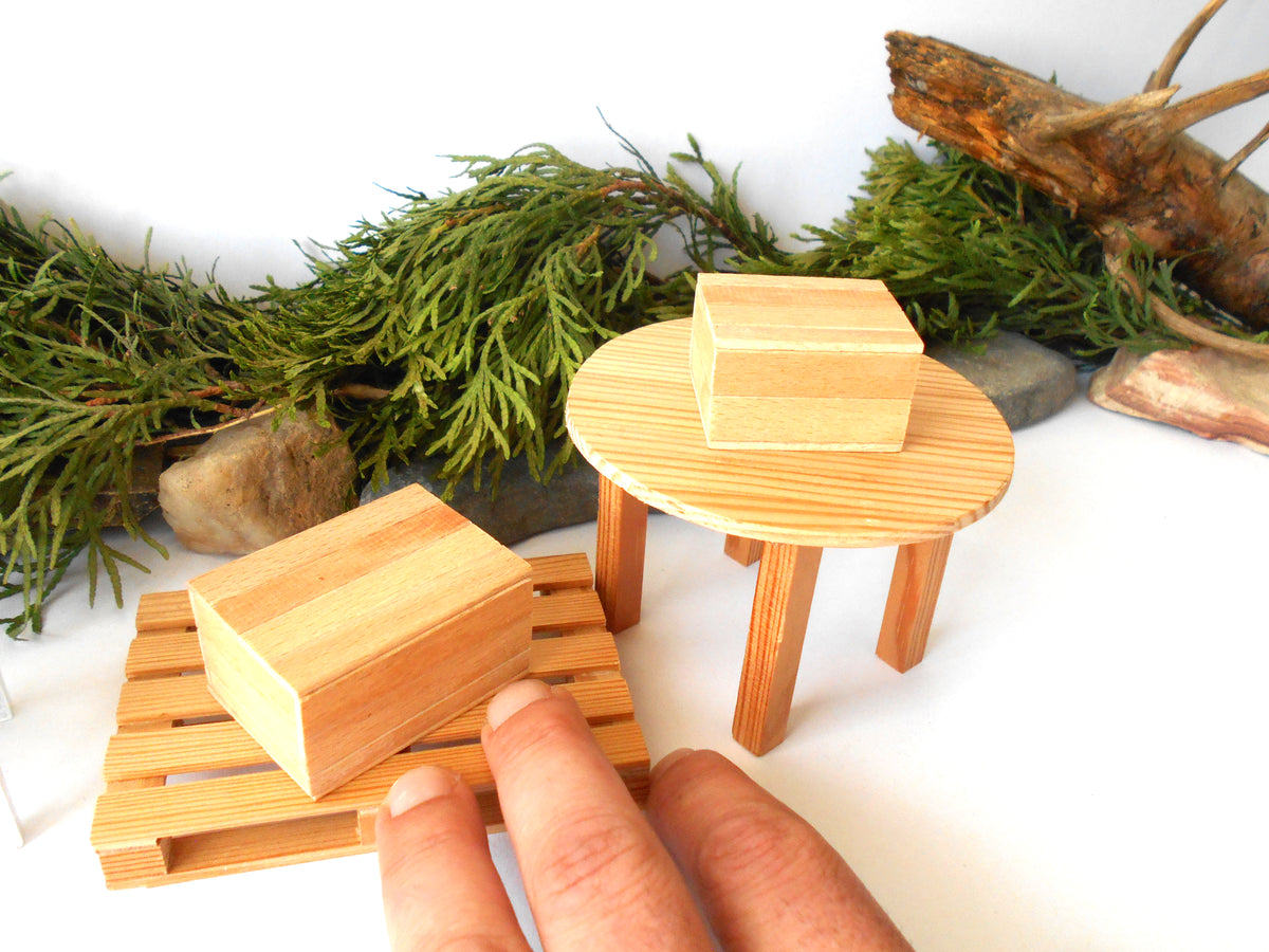 Three small wooden miniature furniture pieces displayed on a white surface with decorative greenery and driftwood in the background. A person's fingers are visible holding one of the wooden blocks, indicating scale.
