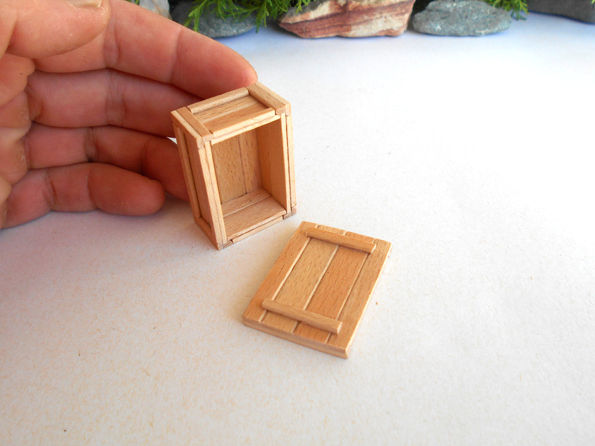 A miniature wooden crate box and its matching lid, shown with a human hand holding the crate to demonstrate scale on a white surface.