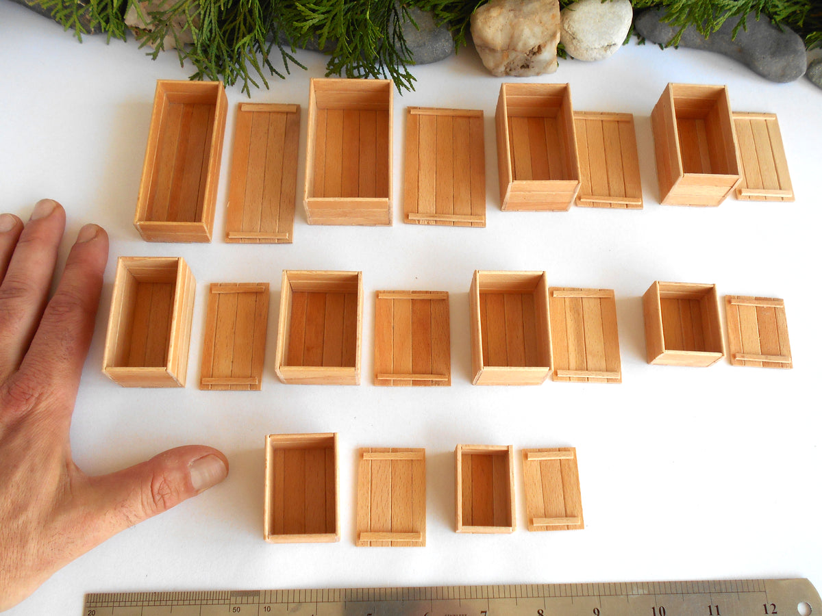 A collection of miniature wooden storage boxes and crates in various sizes, arranged on a white surface with a hand on the left for scale and a ruler at the bottom.
