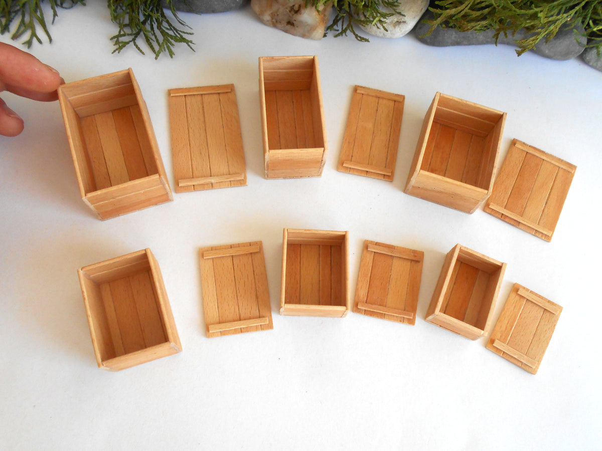 A set of miniature light wood boxes and crates in various sizes, arranged on a white surface with a hand holding one box on the left.