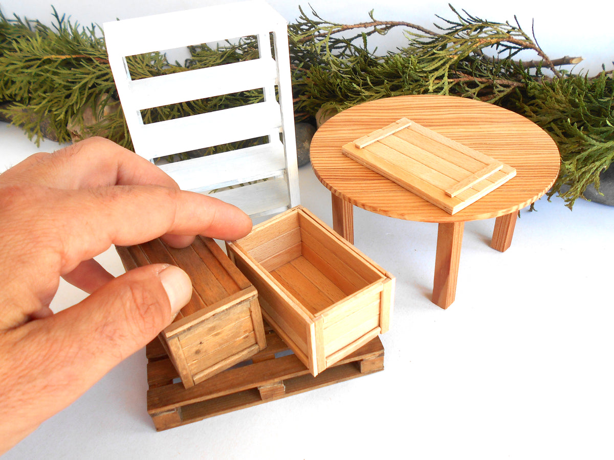 A hand is holding a miniature wooden crate next to a small white chair and round wooden table, with green pine branches in the background.
