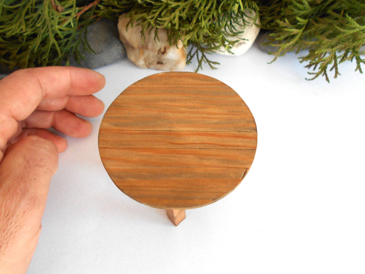 A small round wooden display stand with a natural wood grain finish and a single short leg, shown on a white surface with a hand holding it for scale and greenery in the background.