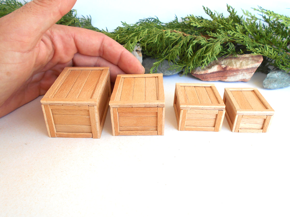 Four miniature light brown wooden crates of different sizes arranged in a row on a white surface, with a person's hand holding the largest crate and green foliage in the background.