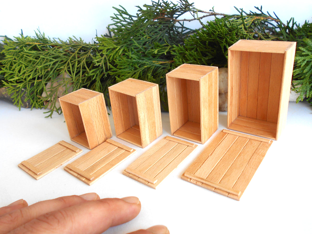A set of miniature wooden box and crate components, including four open storage boxes of varying sizes and four matching slatted lids, displayed on a white surface with green foliage in the background. A person's hand is visible in the bottom-left corner for scale.