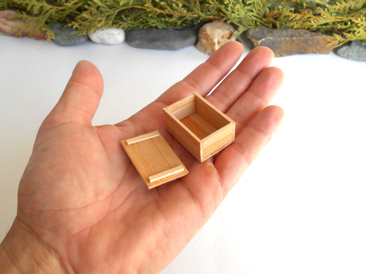 A hand holding two miniature light brown wooden boxes, one closed and one open, with small decorative stones and greenery blurred in the background.