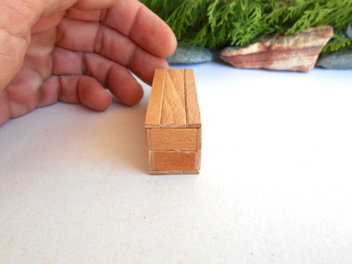 A miniature wooden box with a light brown finish, shown next to a person's hand to demonstrate its small scale, set against a white surface with a blurred green background.