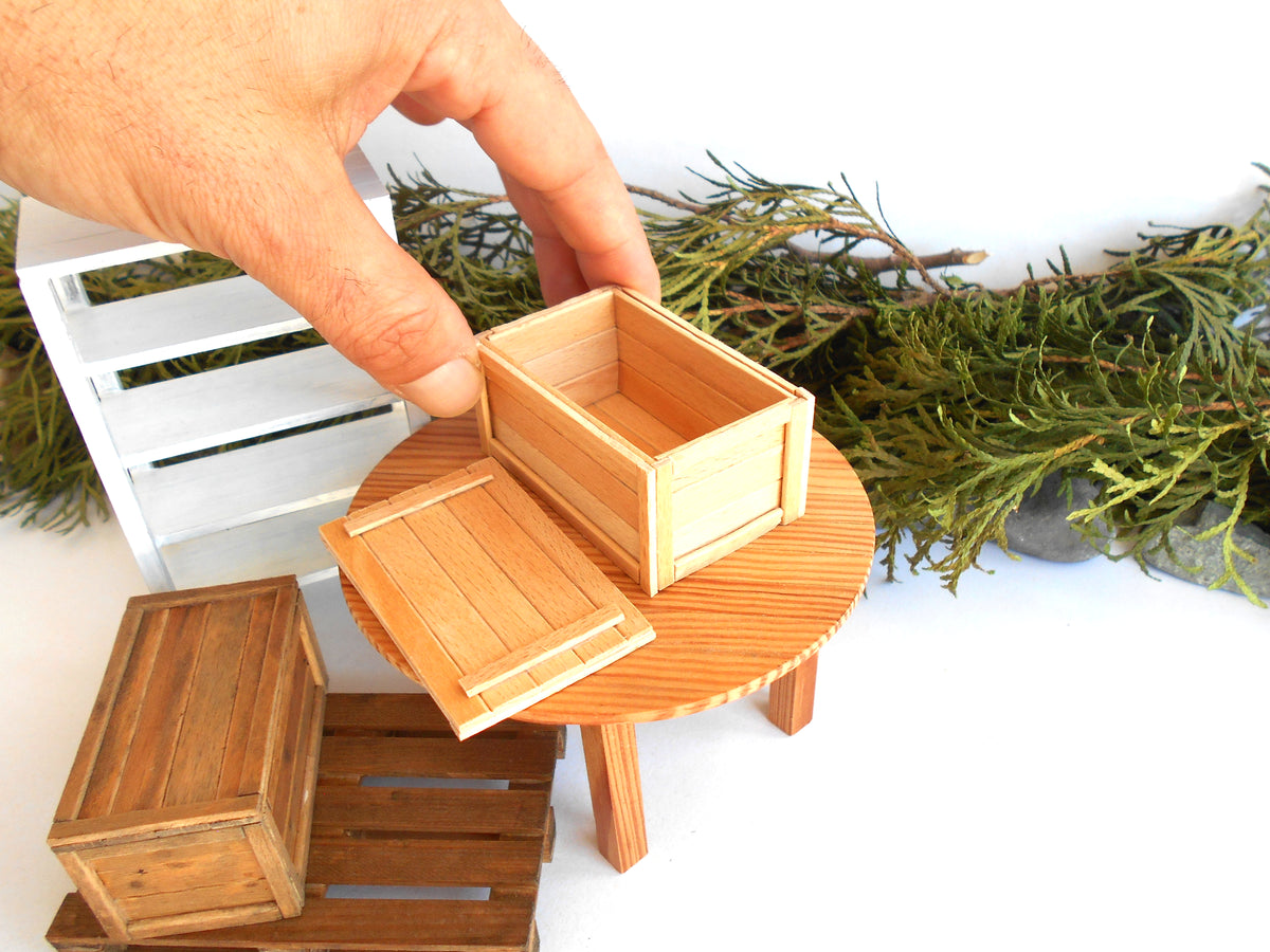 A miniature wooden crate box with its lid off, displayed on a small round wooden table. A human hand is holding the crate, and other miniature wooden crates and green foliage are visible in the background.