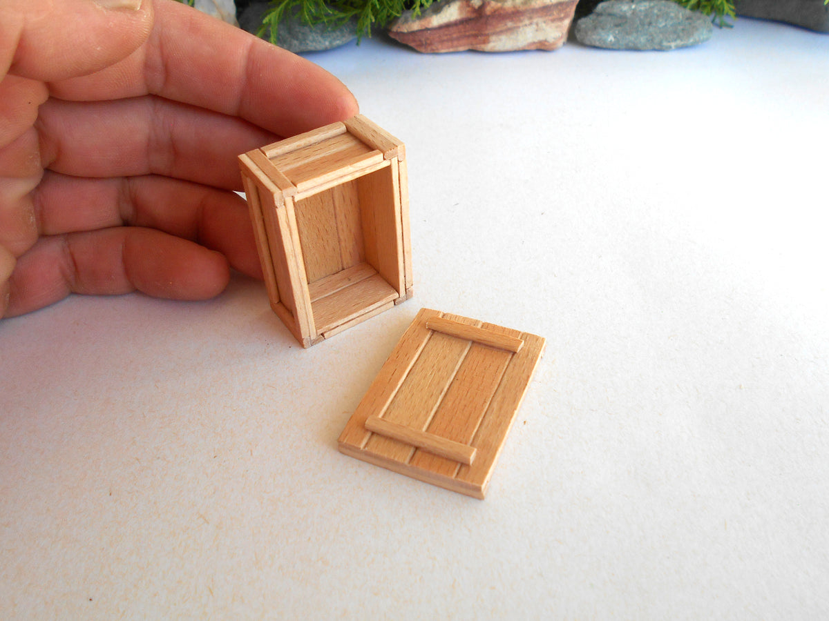 A miniature wooden crate or box with its lid placed beside it, held by a person's hand on a light surface. The product appears to be a small dollhouse or model crate made of light natural wood.