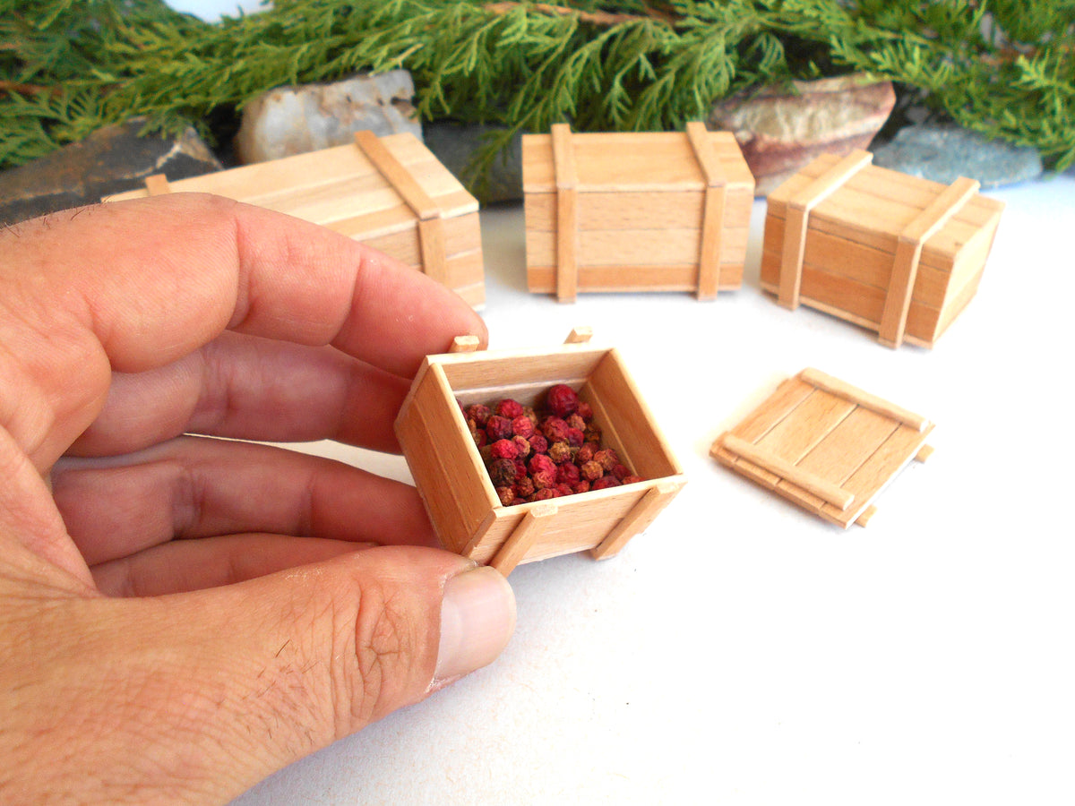 A hand holds a miniature wooden crate filled with small red berries, with several other tiny wooden crates arranged on a white surface in the background.