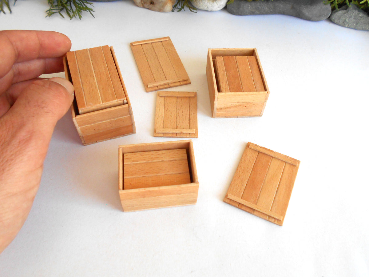 A hand is holding a miniature wooden box lid next to several matching small wooden boxes and lids on a white surface.