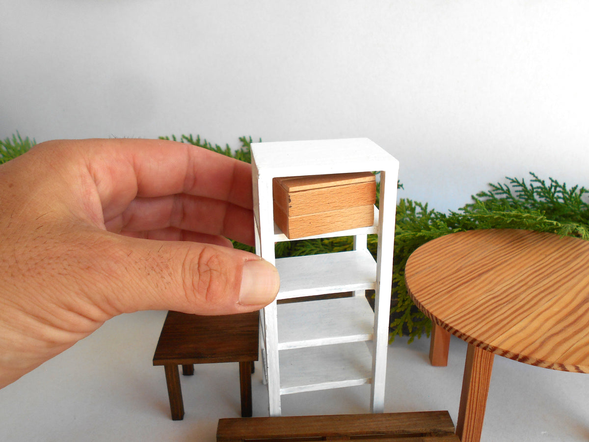 A miniature white shelving unit with a light wood drawer front, held by a human hand to show scale, alongside other miniature furniture pieces.