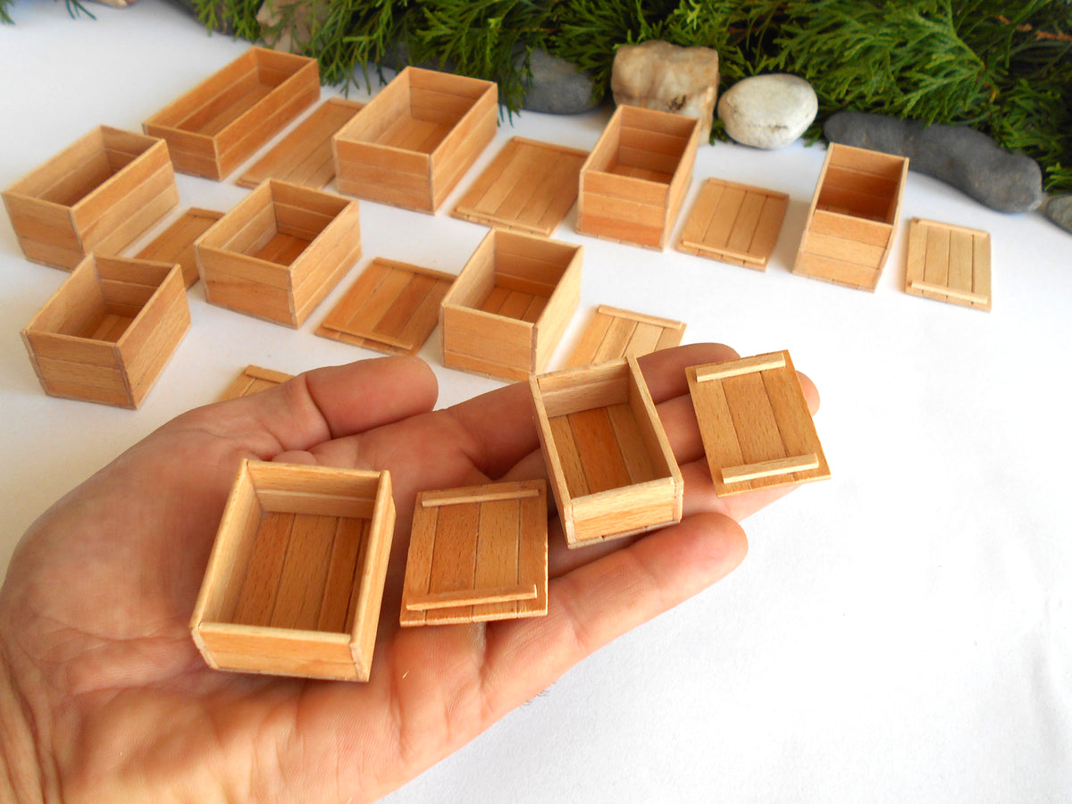 A hand holds several small light wood boxes and lids, with many more of the same arranged on a white surface in the background. The boxes appear to be miniature wooden storage containers or craft boxes.