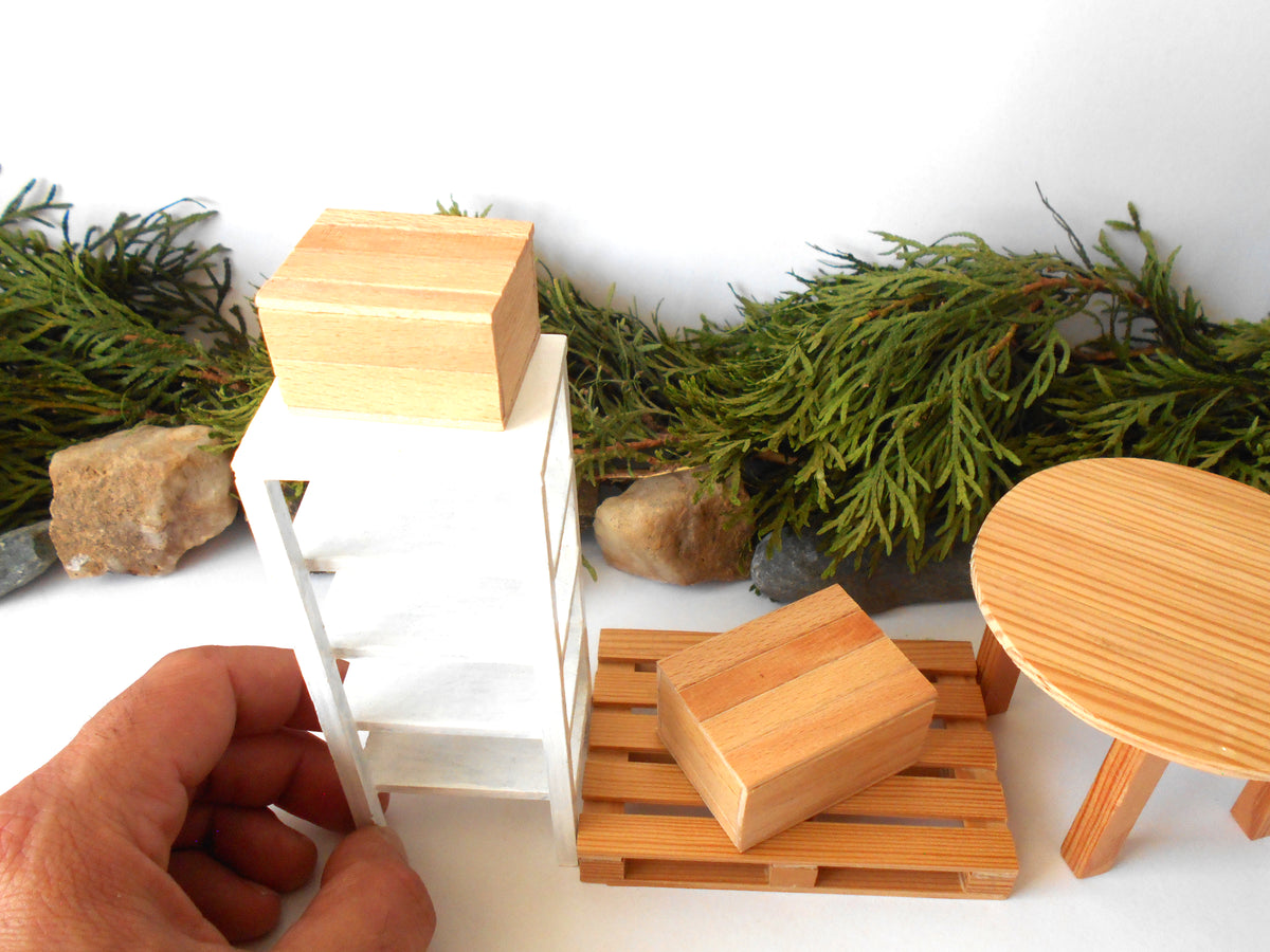 A miniature wooden furniture set displayed on a white surface with green foliage and small rocks in the background. A hand is holding a small white shelving unit on the left, next to a round wooden stool and a pallet-style wooden bench with a wooden block on top.