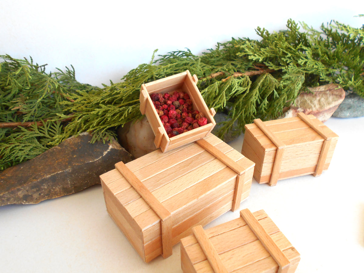 Four miniature light wood crates of different sizes, with one open crate filled with small red berries, arranged on a white surface with green foliage and stones in the background.