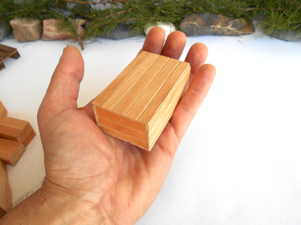 A hand holding a small rectangular wooden block with a slatted top, shown against a white surface with some pine branches and rocks in the background.