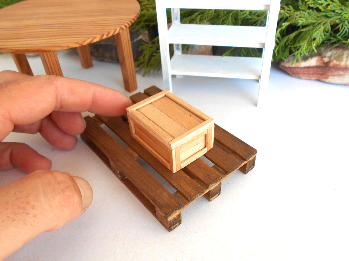 A miniature wooden crate model resting on a small wooden pallet, with a human hand pointing toward it in a close-up shot.