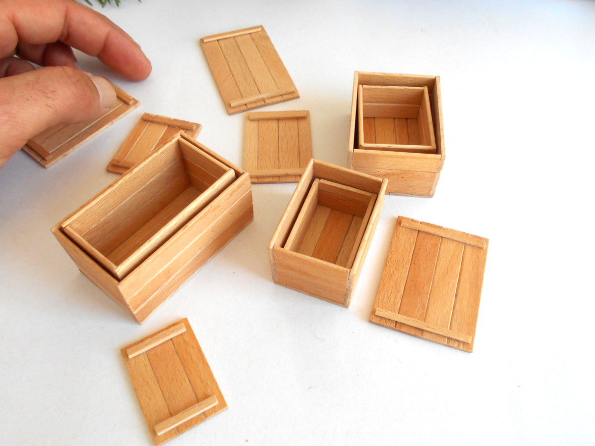 A set of miniature light wood boxes and lids, with a hand visible in the top-left corner interacting with one of the boxes, all arranged on a plain white surface.