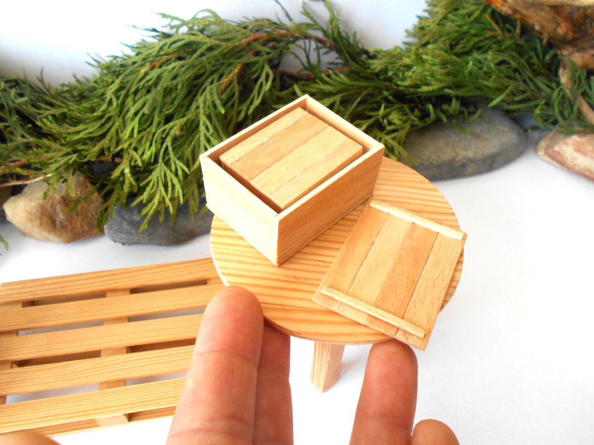 A miniature light wood dollhouse accessory set, including a small wooden box, a round stand, and a slatted base, held by a person's hand against a white background with decorative greenery.