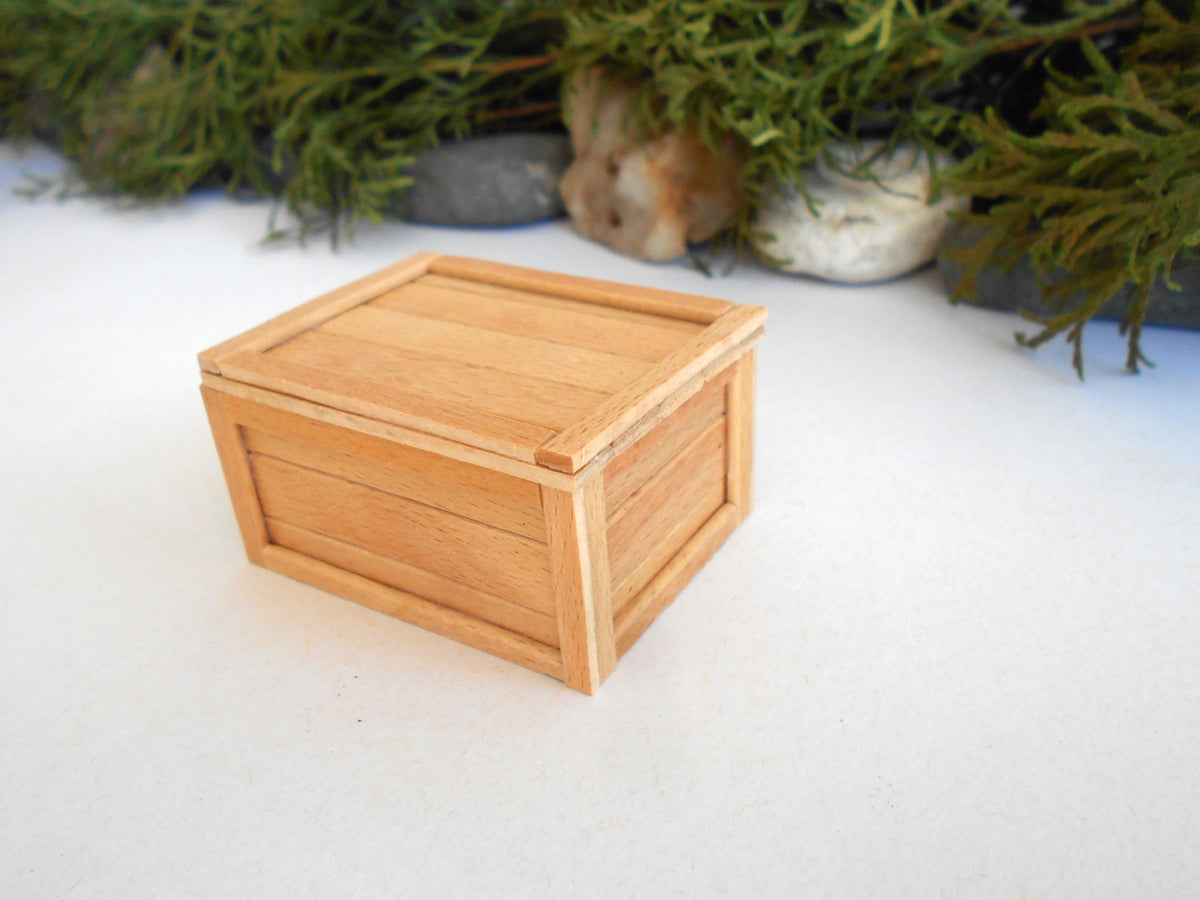 A small square wooden box with a slatted lid, shown on a white surface with blurred greenery and stones in the background.