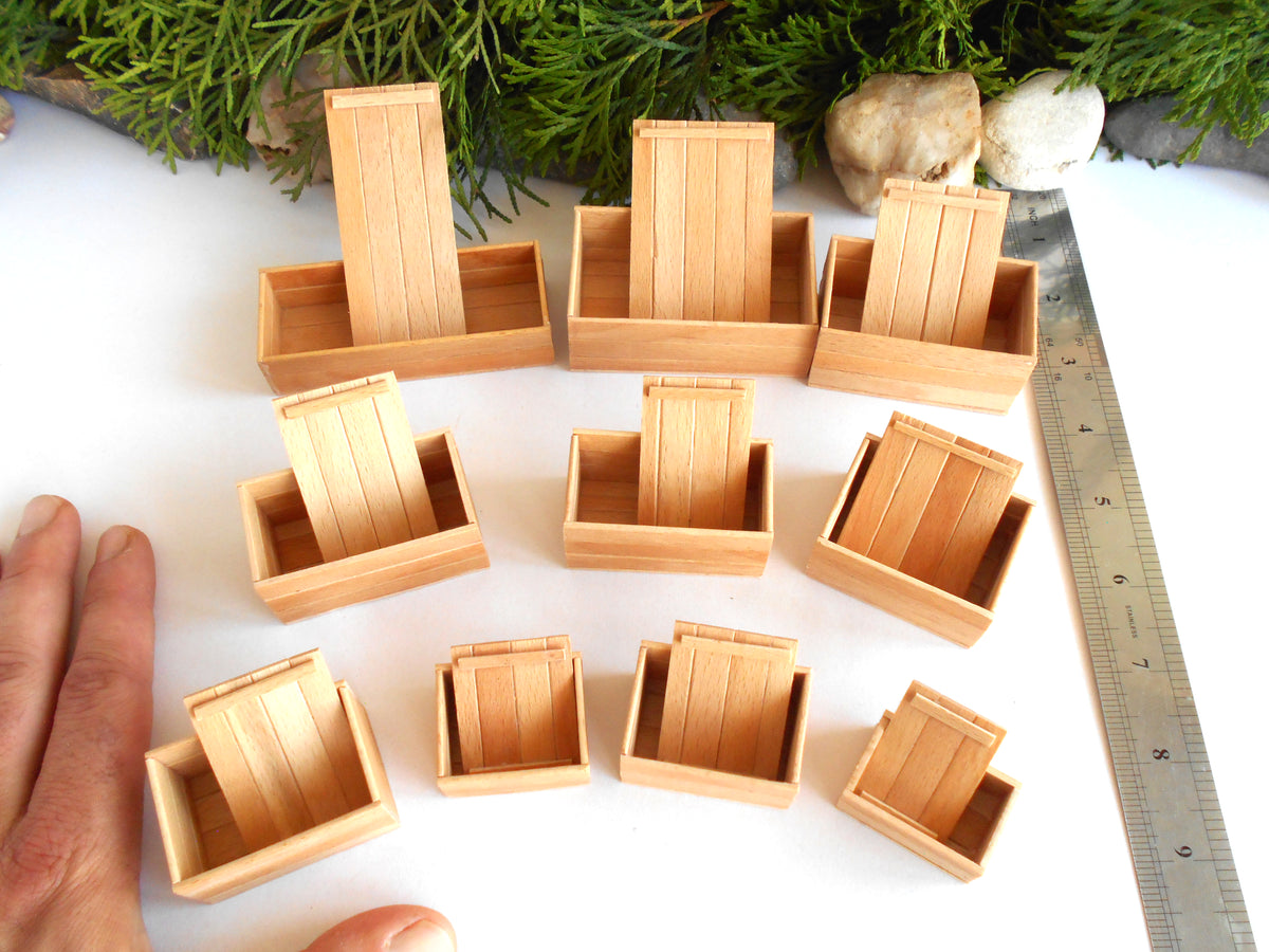 Ten miniature light wood garden planters arranged on a white surface, with a hand visible at the bottom left for scale and a metal ruler on the right. Green foliage and small stones decorate the background.