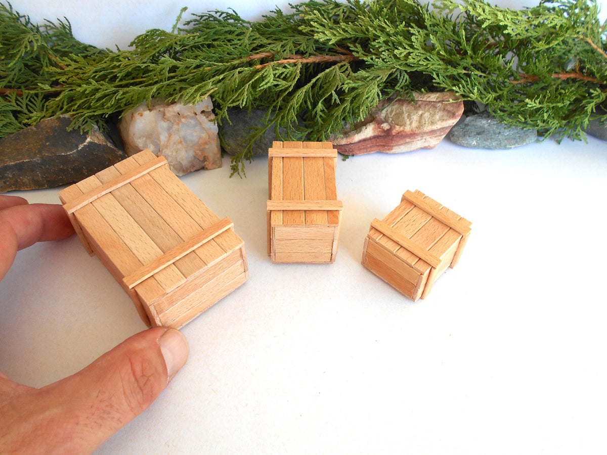 Three miniature light wood crates of different sizes displayed on a white surface, with a hand holding the largest crate for scale and a green pine garland with stones in the background.