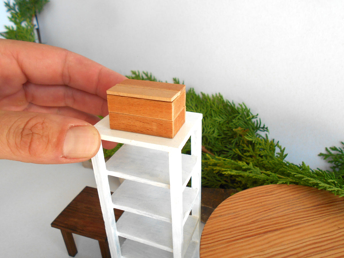 A miniature wooden box with a light brown finish is being held by a hand on a white miniature shelving unit. The scene is set against a white wall with green foliage and a round wooden table in the background, suggesting a dollhouse or miniature scale model context.