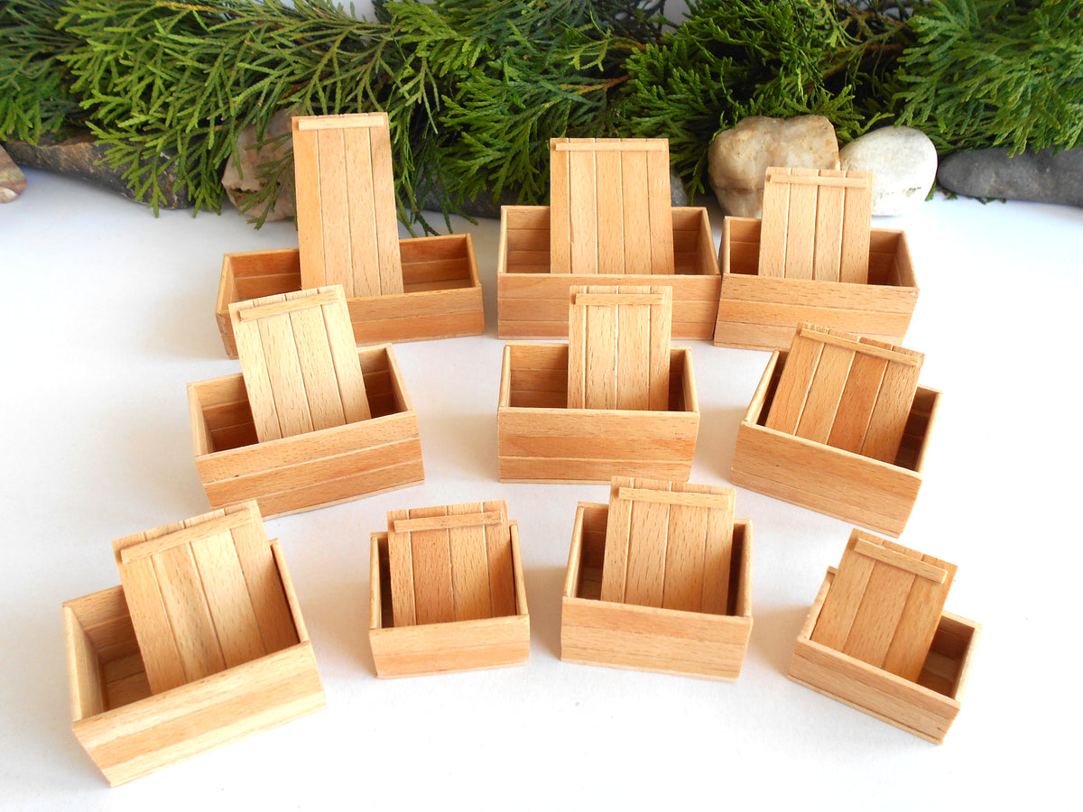 A set of miniature wooden crate dollhouse accessories, shown in various open and closed positions on a white surface with green pine branches and small stones in the background.