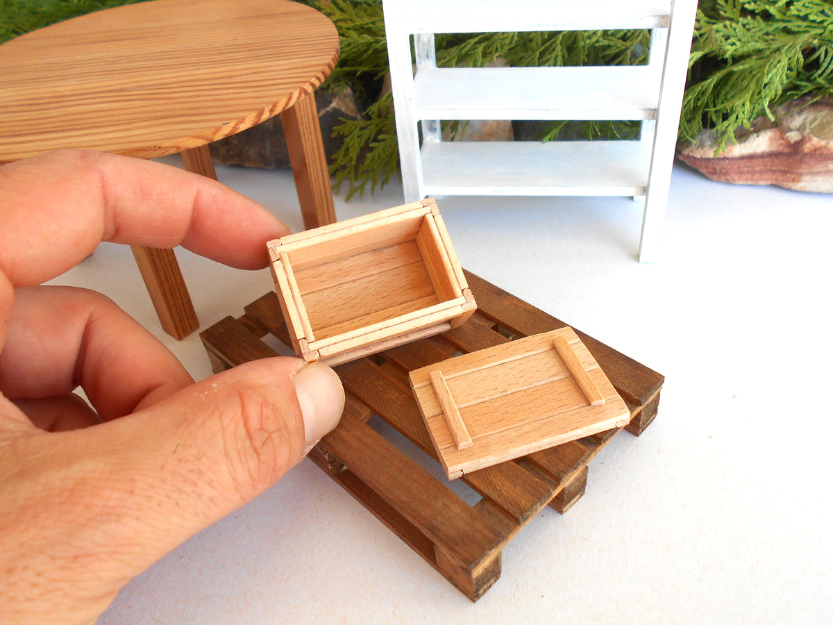 A close-up of a hand holding miniature wooden crates, with a small wooden pallet base and miniature furniture pieces in the background.
