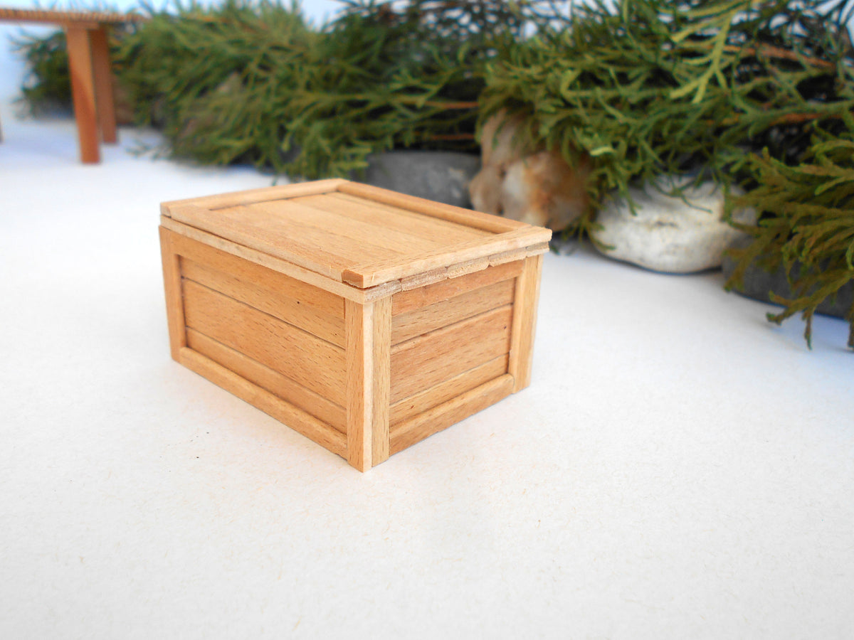 A miniature light wood crate box with a slatted lid, shown on a white surface with a blurred background of green foliage.