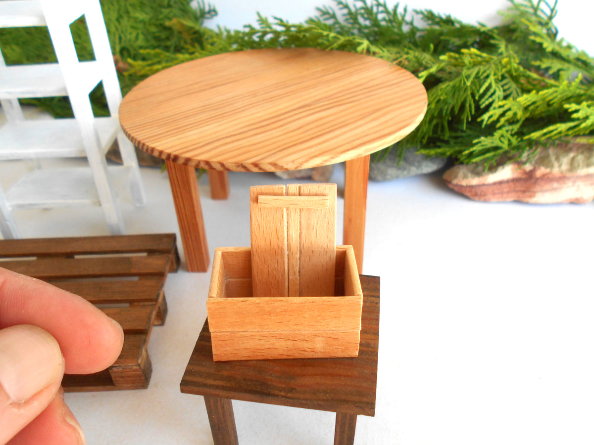 A miniature wooden box with a slatted front and back, displayed on a small dark wooden stand. A human hand is visible in the bottom-left corner holding the scale, with a miniature round table, white chair, and green foliage in the background.