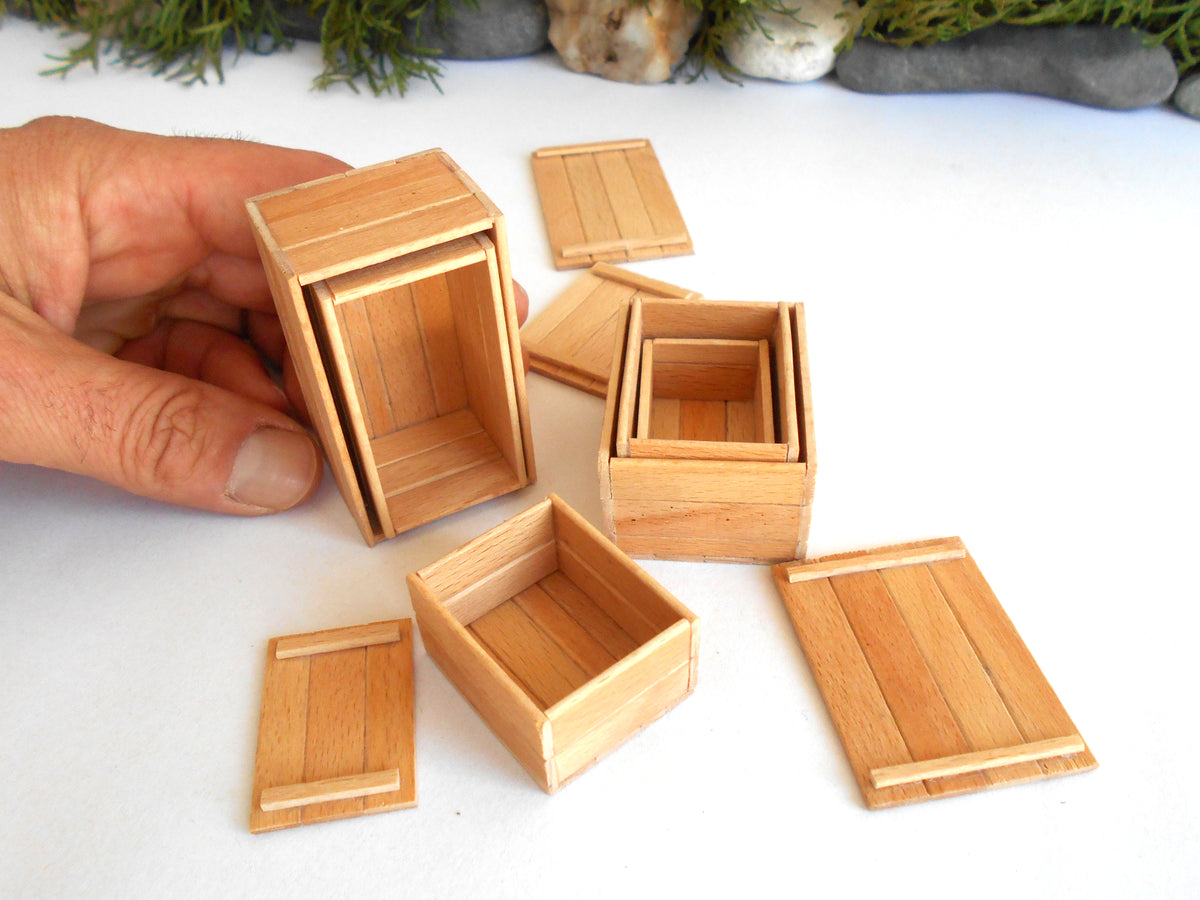 A hand holding a miniature wooden box, with several small wooden storage boxes and lids arranged on a white surface. The boxes appear to be doll-scale or tiny wooden containers with visible grain and simple construction.