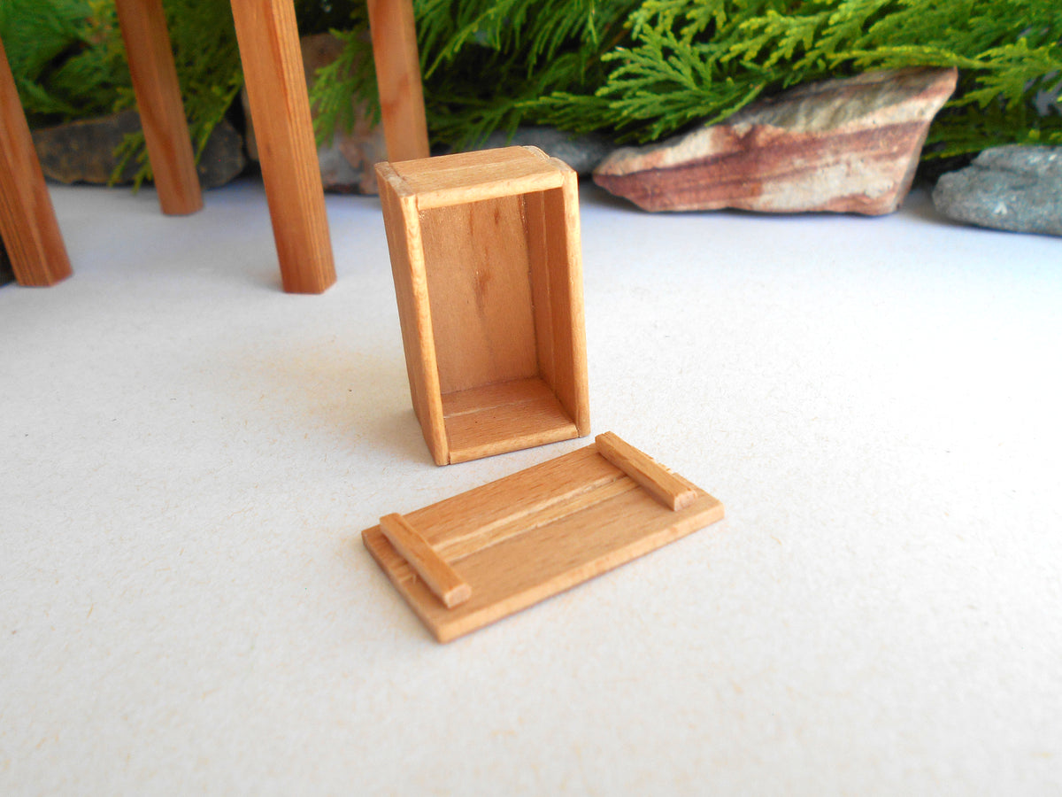 A miniature light brown wooden box with its lid placed in front, set on a white surface with a blurred background of green foliage and rocks.