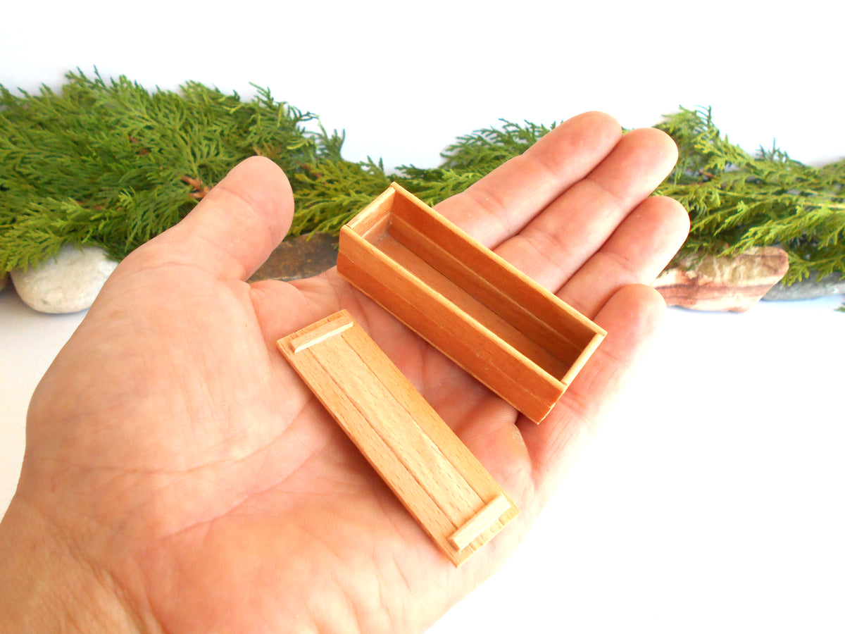 A miniature light brown wooden box and its matching lid held in a person's hand, with green moss and small stones in the background.