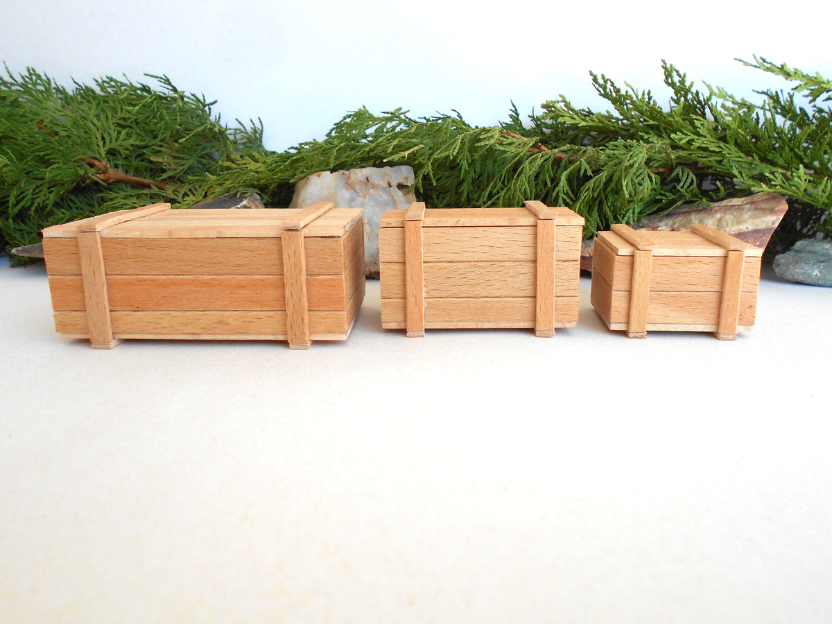 Three miniature wooden crates of different sizes arranged in a row on a light beige surface, with green pine branches and small stones in the background.