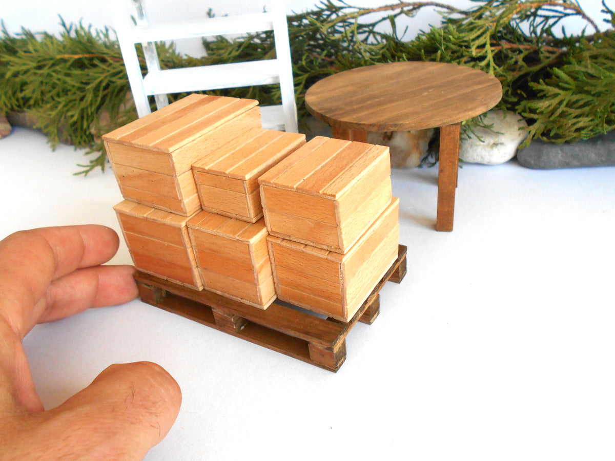 A miniature wooden pallet stacked with small rectangular wood miniature boxes , shown next to a human hand for scale, with a miniature round table and greenery in the background.