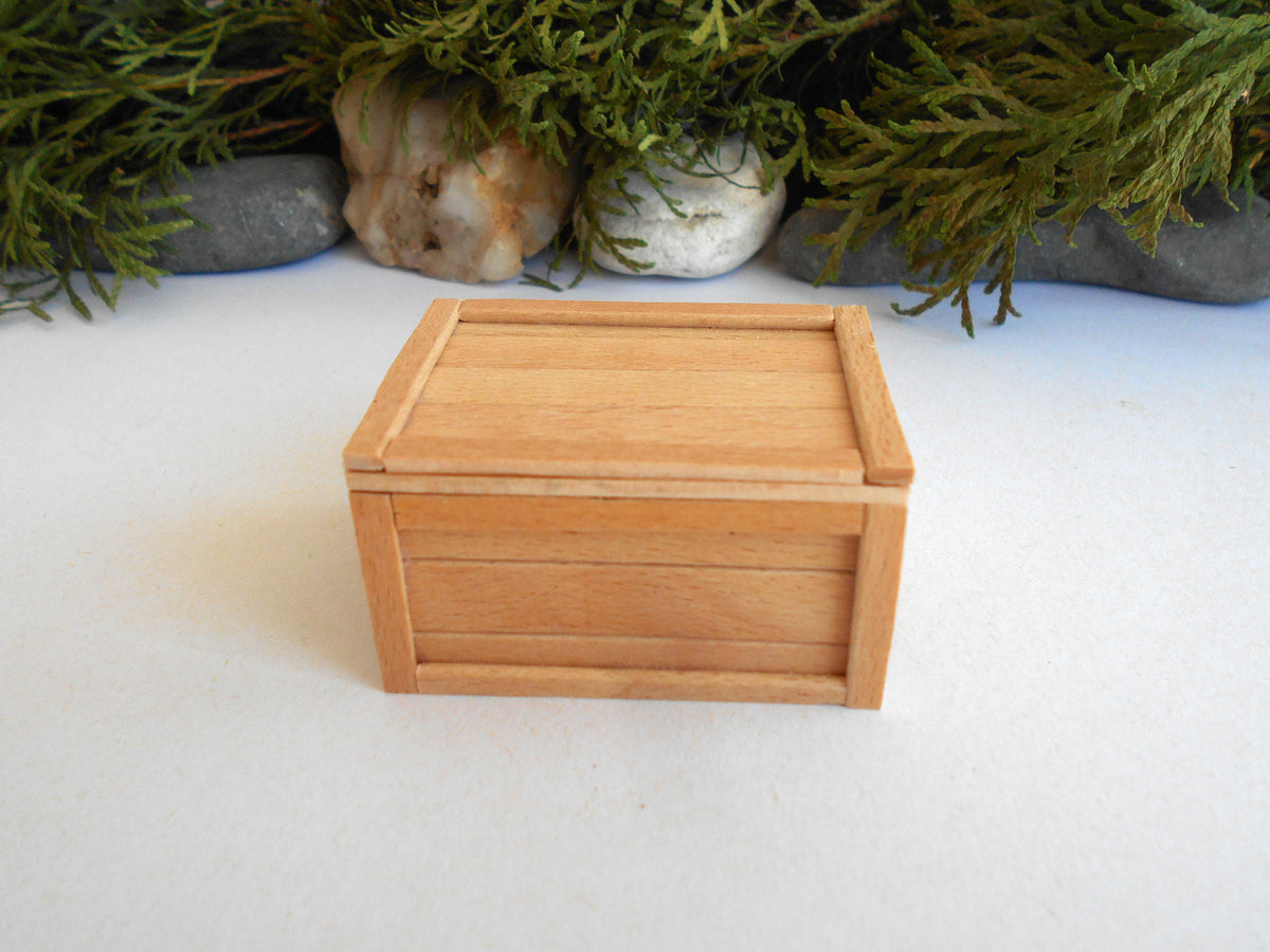 A miniature light brown wooden box with a hinged lid, displayed on a white surface with a blurred background of green foliage and stones.