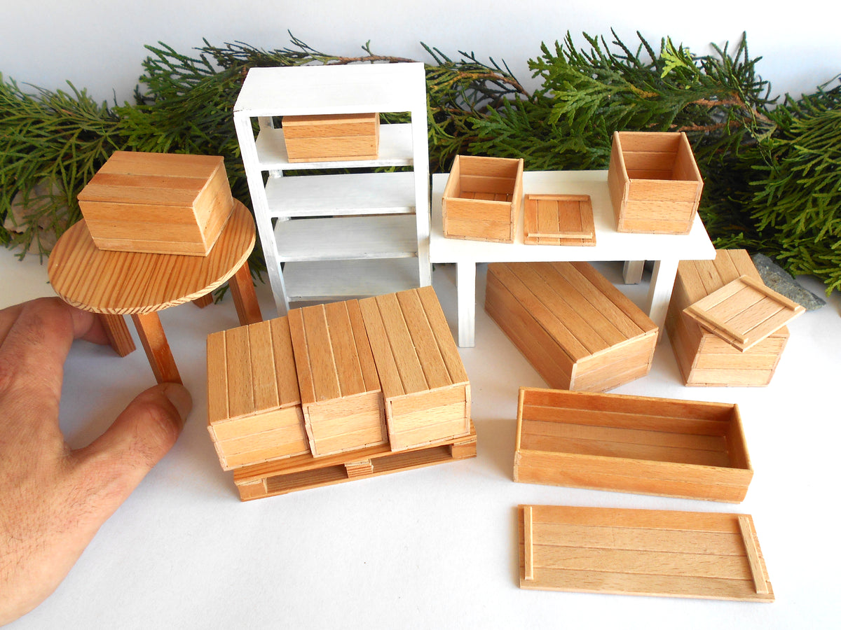 A miniature dollhouse furniture set featuring light wood crates, shelves, and tables, with a white table and shelving unit, displayed on a white surface with green foliage in the background. A human hand is visible on the left holding one of the wooden crates to show scale.