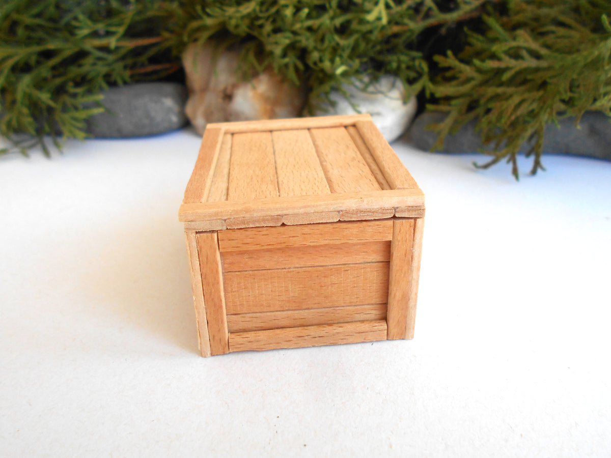 A miniature light brown wooden box with a slatted lid, photographed on a white surface with blurred green foliage and gray stones in the background.