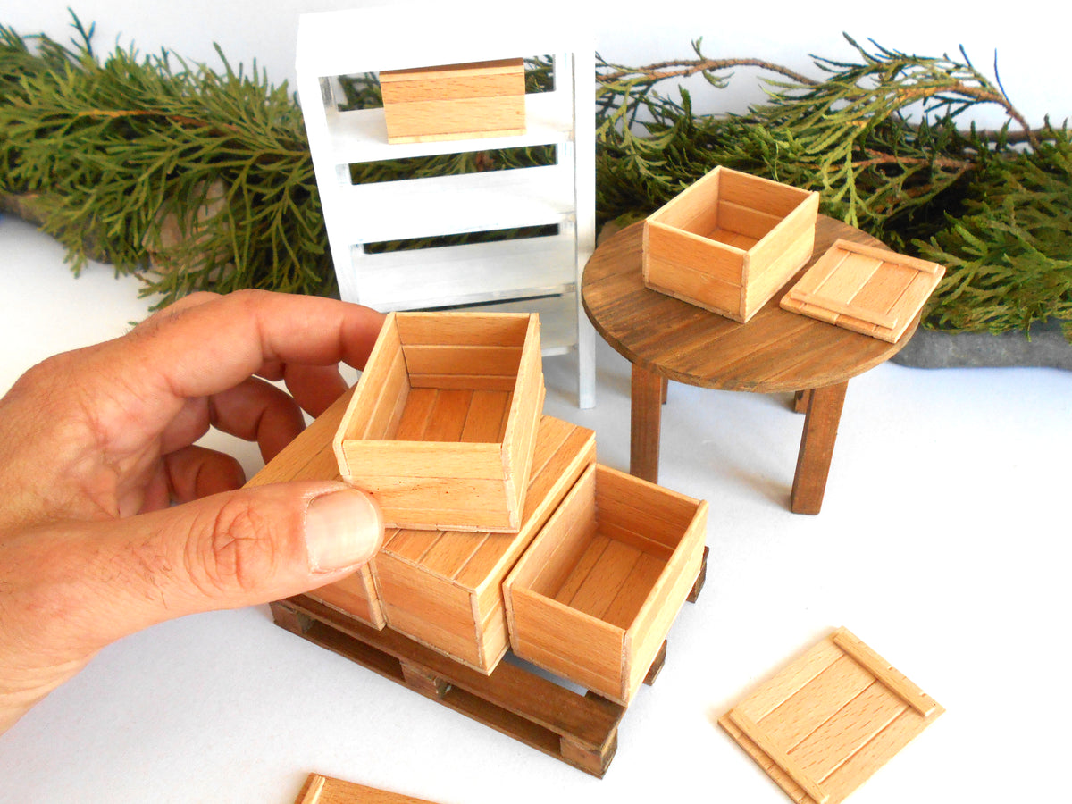 A hand holds a miniature wooden crate in a dollhouse-scale setting, with additional small wooden storage boxes, a round table, shelving, and greenery in the background.