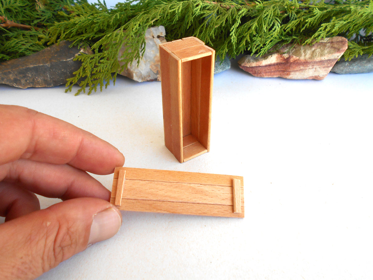 A hand holds a small rectangular wooden box lid in front of a white surface, with another matching open wooden box standing upright in the background. Greenery and stones are visible behind the setup.