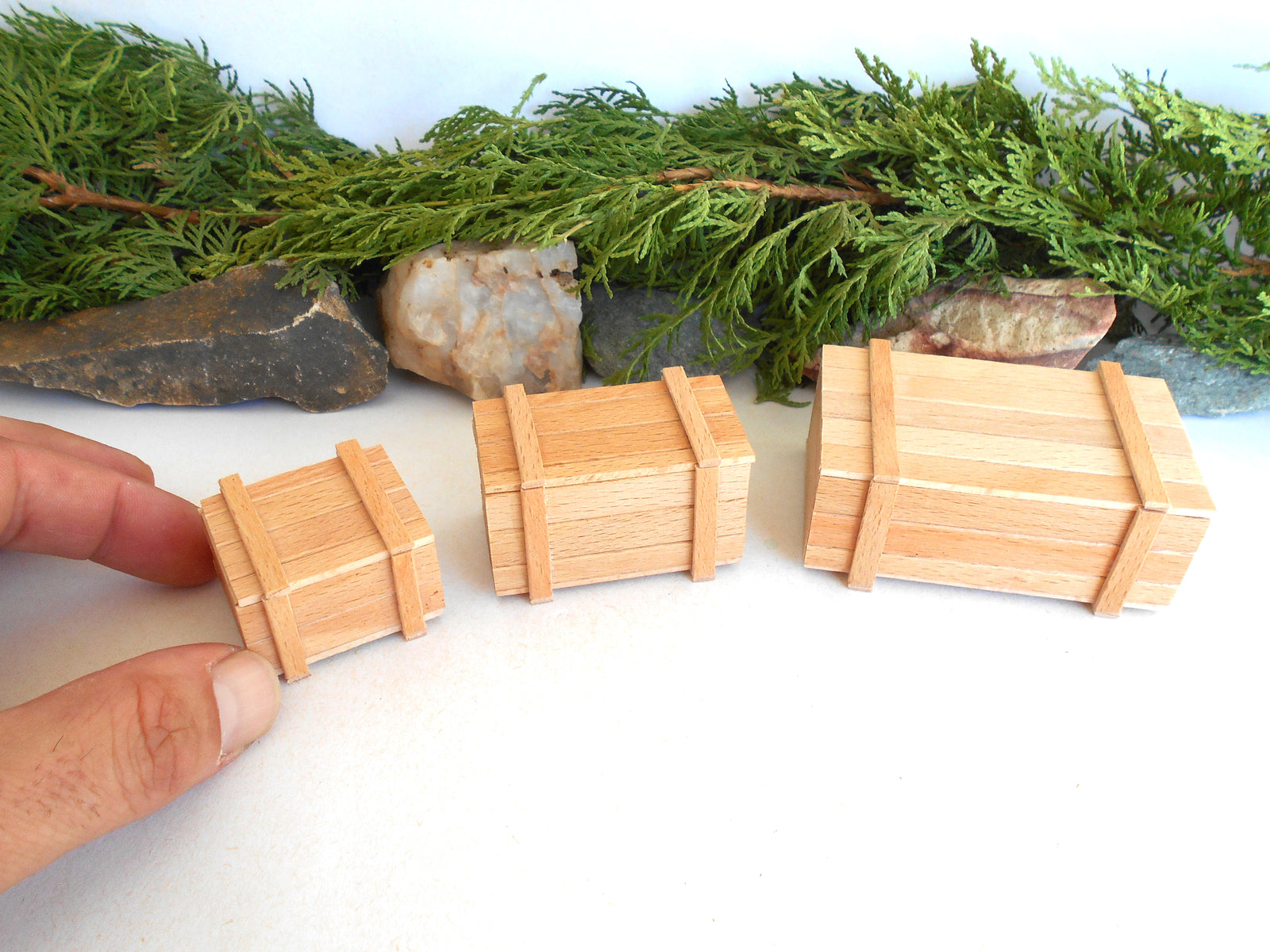 Three miniature wooden crates of different sizes displayed on a white surface with green foliage and rocks in the background. A hand is holding the smallest crate on the left.